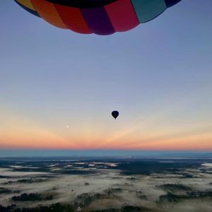 Fog on the ground with orange, yellow, and blue sun rays next to a hot air balloon flying over Lake Tapps